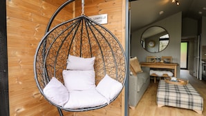 Una habitación con una hamaca colgante, un muro de madera, una zona de comedor con una mesa y sillas, y una cocina visible a través de una puerta abierta.