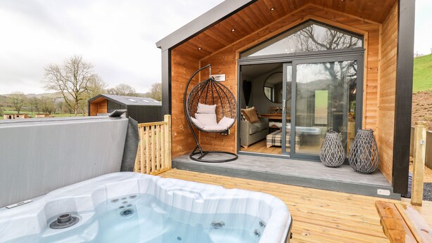 Un área moderna de hidromasaje al aire libre con un deck de madera, una silla colgante y un ventanal de vidrio que da a un espacio interior acogedor.
