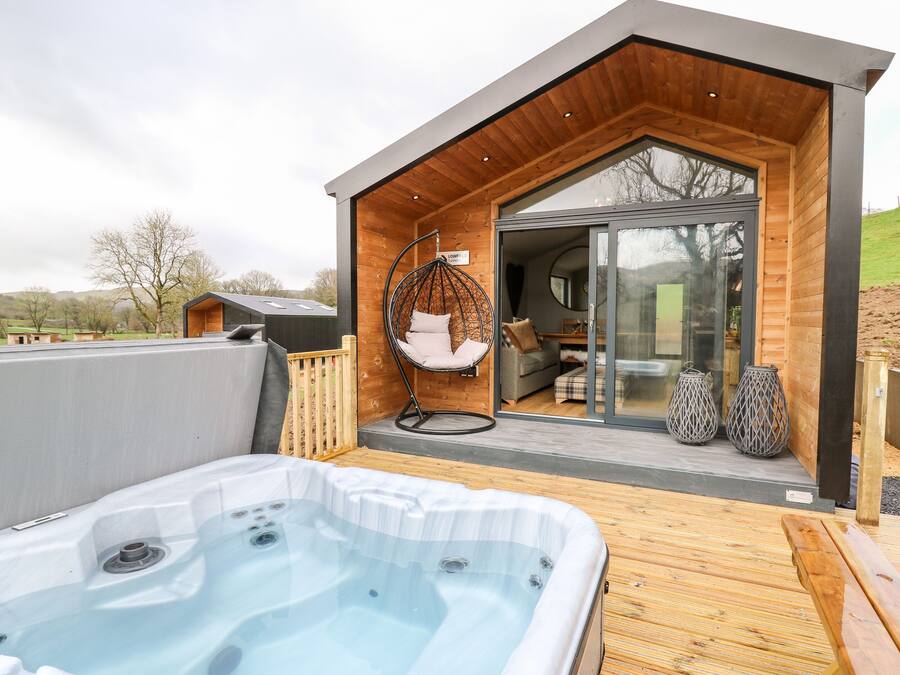A modern outdoor hot tub area with a wooden deck, a hanging chair, and a glass door leading to a cozy interior space.
