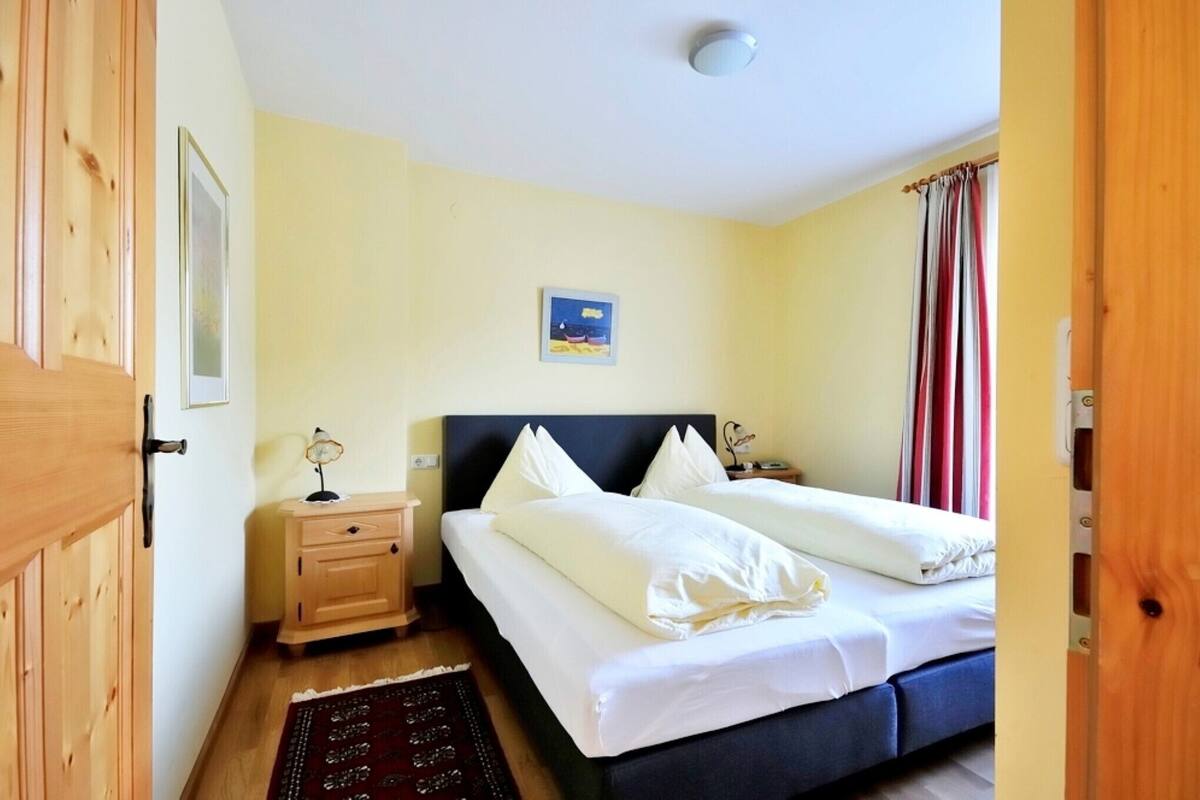 A bedroom with two beds, a bedside table with a lamp, a framed picture on the wall, and a window with curtains.