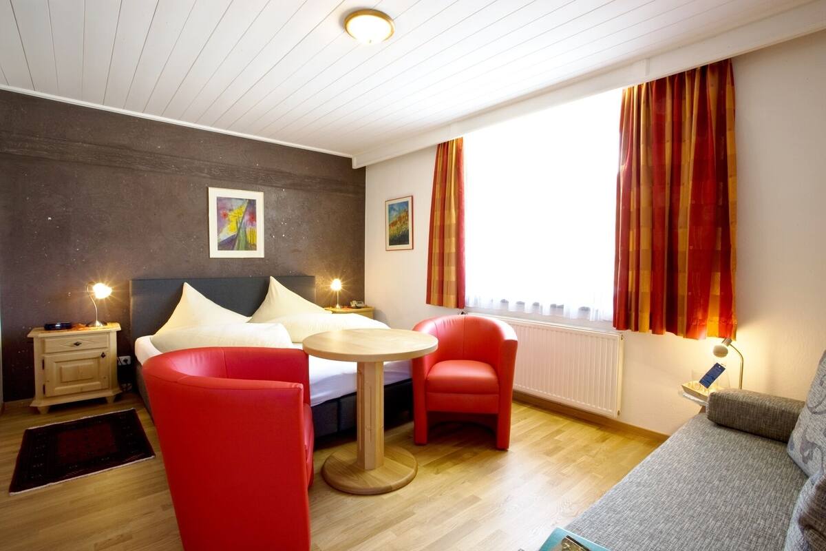 A modern hotel room with a bed, a round table, two red chairs, a grey sofa, and a window with curtains.