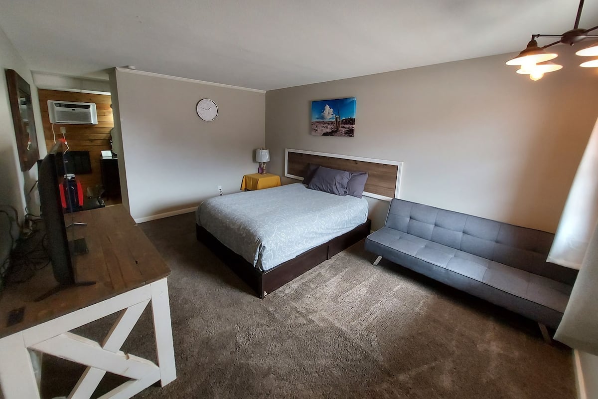 A bedroom with a bed, a sofa, a desk, a lamp, a clock, and a painting on the wall.