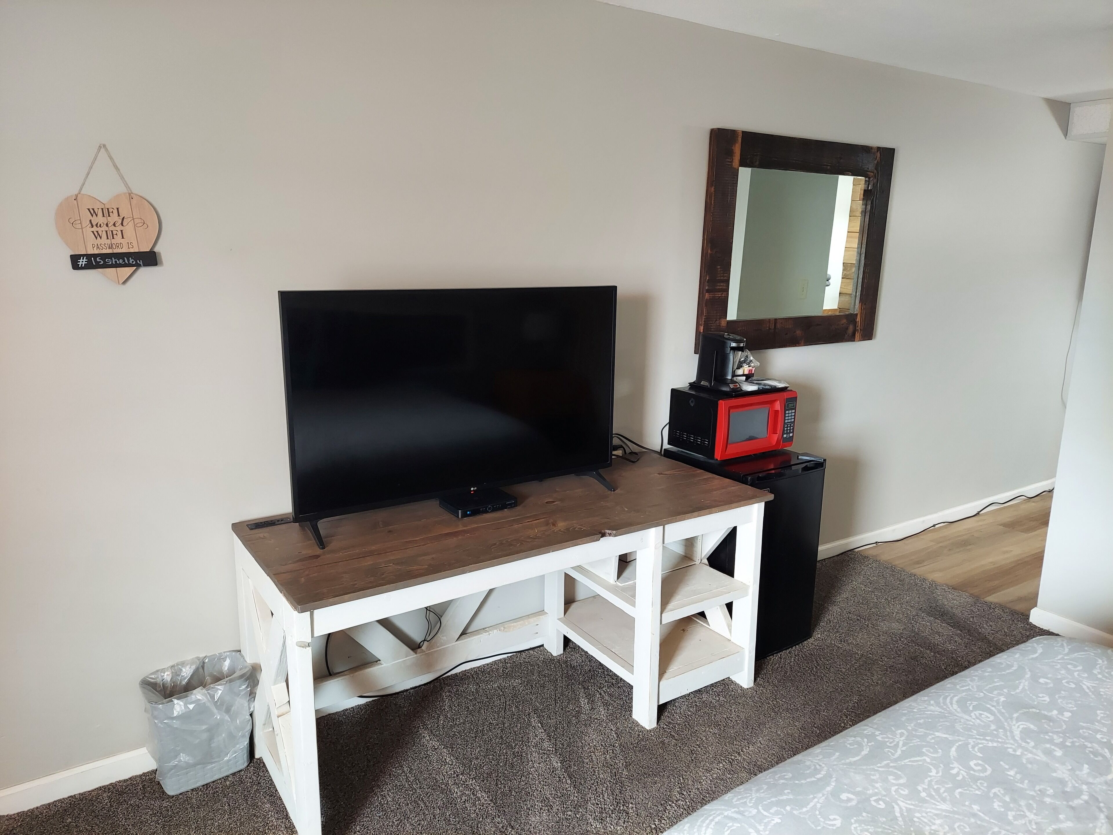 Una habitación pequeña con una televisión sobre una mesa de madera, un microondas rojo, un cesto de basura y un espejo en la pared.