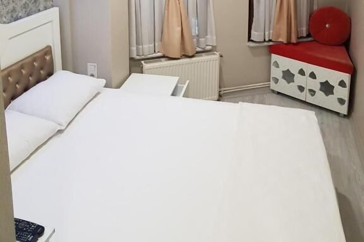 A hotel room with a bed, a red cushioned bench, a radiator, and curtains.