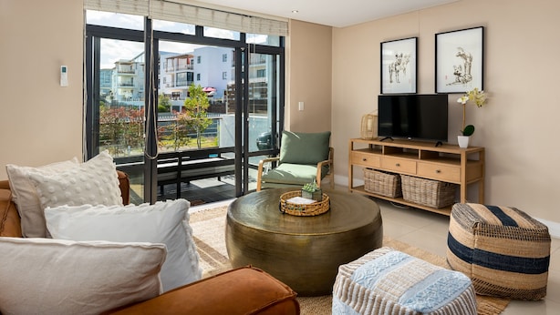 Een moderne woonkamer met een bank, voetenbankje en een salontafel. Er staat een televisie op een houten dressoir en een groot raam met uitzicht op gebouwen.
