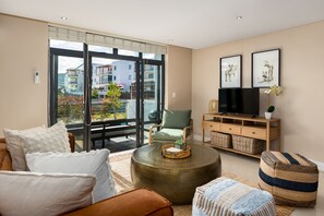 A modern living room with a sofa, ottoman, and a coffee table. There is a television on a wooden stand and a large window with a view of buildings. - Eden On The Bay Apartment | Beachfront | Netflix (Cape Town)