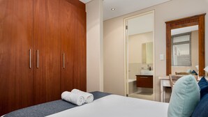 Hotelkamer met een grote houten garderobe, een bed met witte lakens, een badkamer met wastafel en spiegel, en een raam met luxaflex.