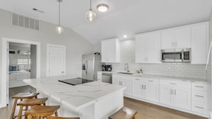 Una cocina moderna con armarios blancos, una isla grande con taburetes, electrodomésticos de acero inoxidable e iluminación colgante.