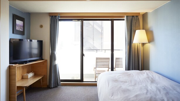 A hotel room with a bed, a TV, a desk, a lamp, and a sliding glass door to a balcony.
