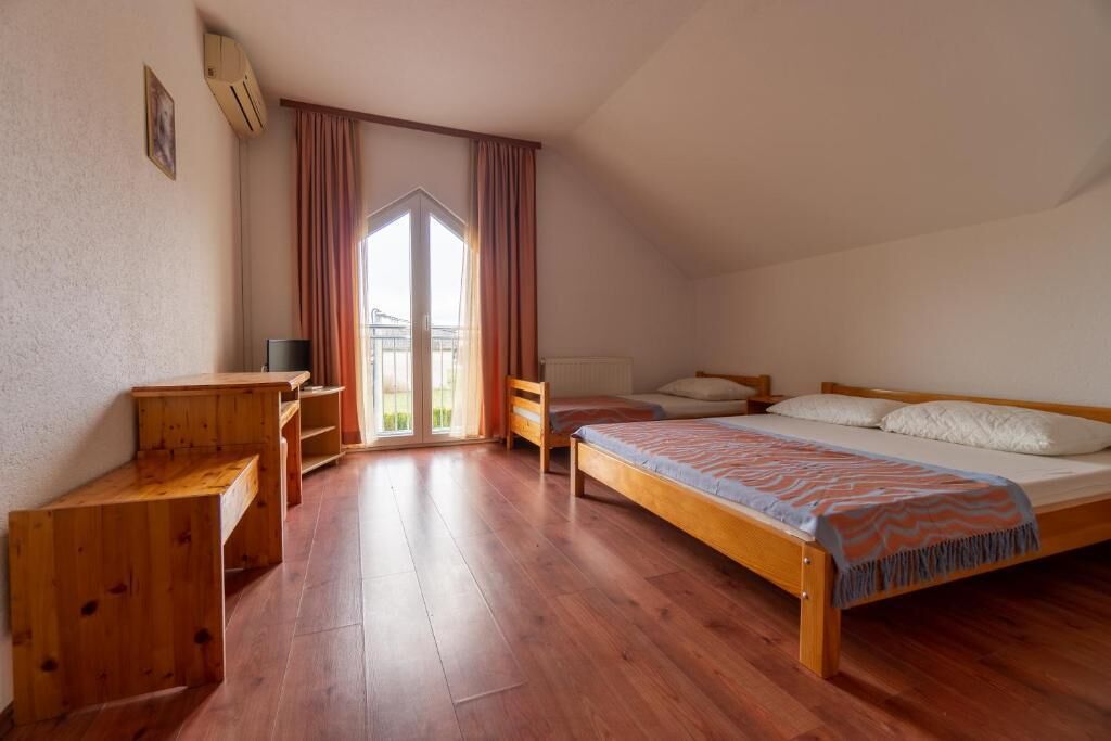 Een slaapkamer met een houten bed, een bureau en een stoel. Er is een raam met gordijnen, en een radiator aan de muur.