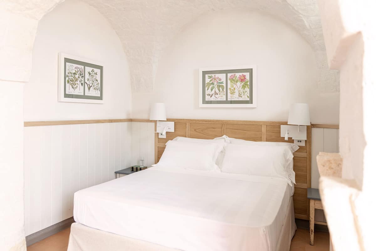 Quarto com cabeceira de madeira, roupa de cama branca, duas luminárias fixadas na parede e duas ilustrações botânicas emolduradas.