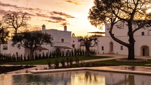 Une cour avec une piscine, des bâtiments blancs et des arbres, le tout baigné par les couleurs du coucher de soleil.