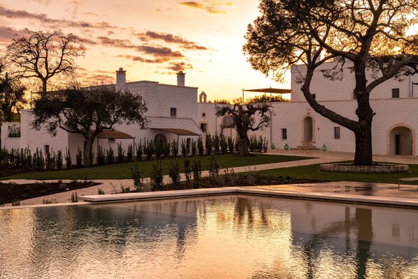 Un patio con piscina, edificios blancos y árboles al atardecer.