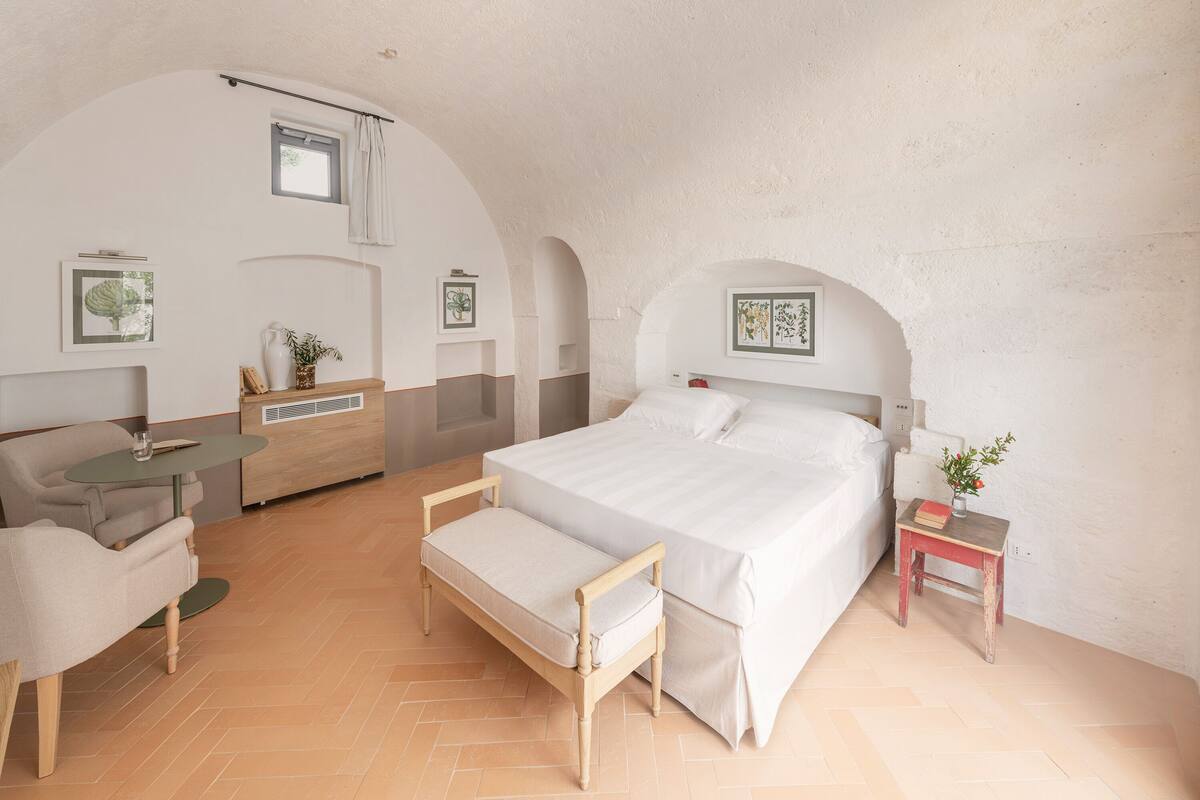 Um quarto com cama, mesa de cabeceira, cadeira, uma pequena mesa e um sofá.