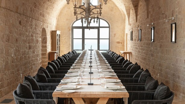 Une longue table à manger, entourée de chaises, se trouve dans une pièce aux murs en pierre et au plafond haut.