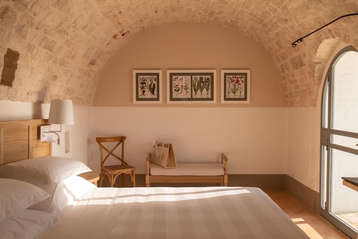 Quarto com cama e cadeira de madeira, um banco e ilustrações botânicas emolduradas na parede.