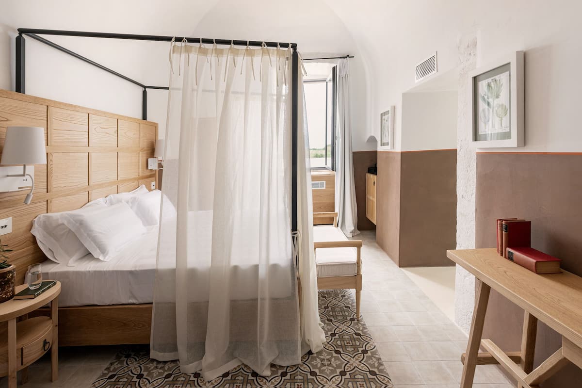 Quarto com cabeceira de madeira, cama com lençóis brancos, mesa de cabeceira com luminária, planta na mesa de cabeceira e janela com cortinas leves.