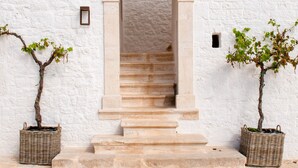 Un bâtiment blanc avec un escalier en pierre menant à une entrée en arc de cercle. De part et d’autre de l’entrée, on trouve deux plantes en pot.