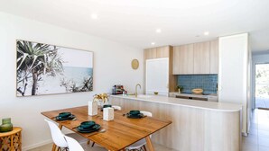 A modern kitchen with a wooden dining table, white chairs, and a view of the sea through a window. - Exquisite 1-Bed Apartment with Bay Views (Batemans Bay)