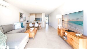 A modern living room with a sofa, dining table, and TV. A kitchen is visible in the background. - Exquisite 1-Bed Apartment with Bay Views (Batemans Bay)