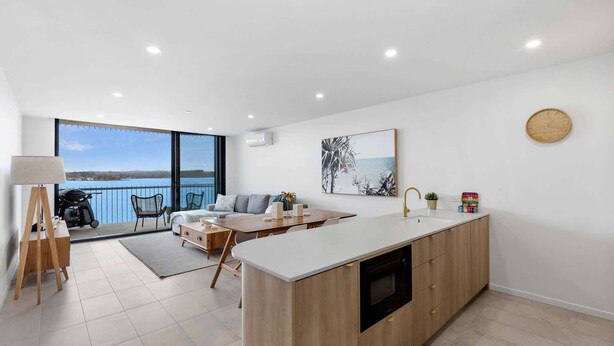 A modern kitchen with a large island, a dining area with a view of the sea, and a balcony with a view of the water. - Exquisite 1-Bed Apartment with Bay Views (Batemans Bay)