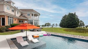 A swimming pool with lounge chairs and colorful inflatable rings, a large umbrella, and a house in the background. - 6BR Waterfront Estate | Private Dock, Firepit, Screened Porch & Beach Access (Sherwood)
