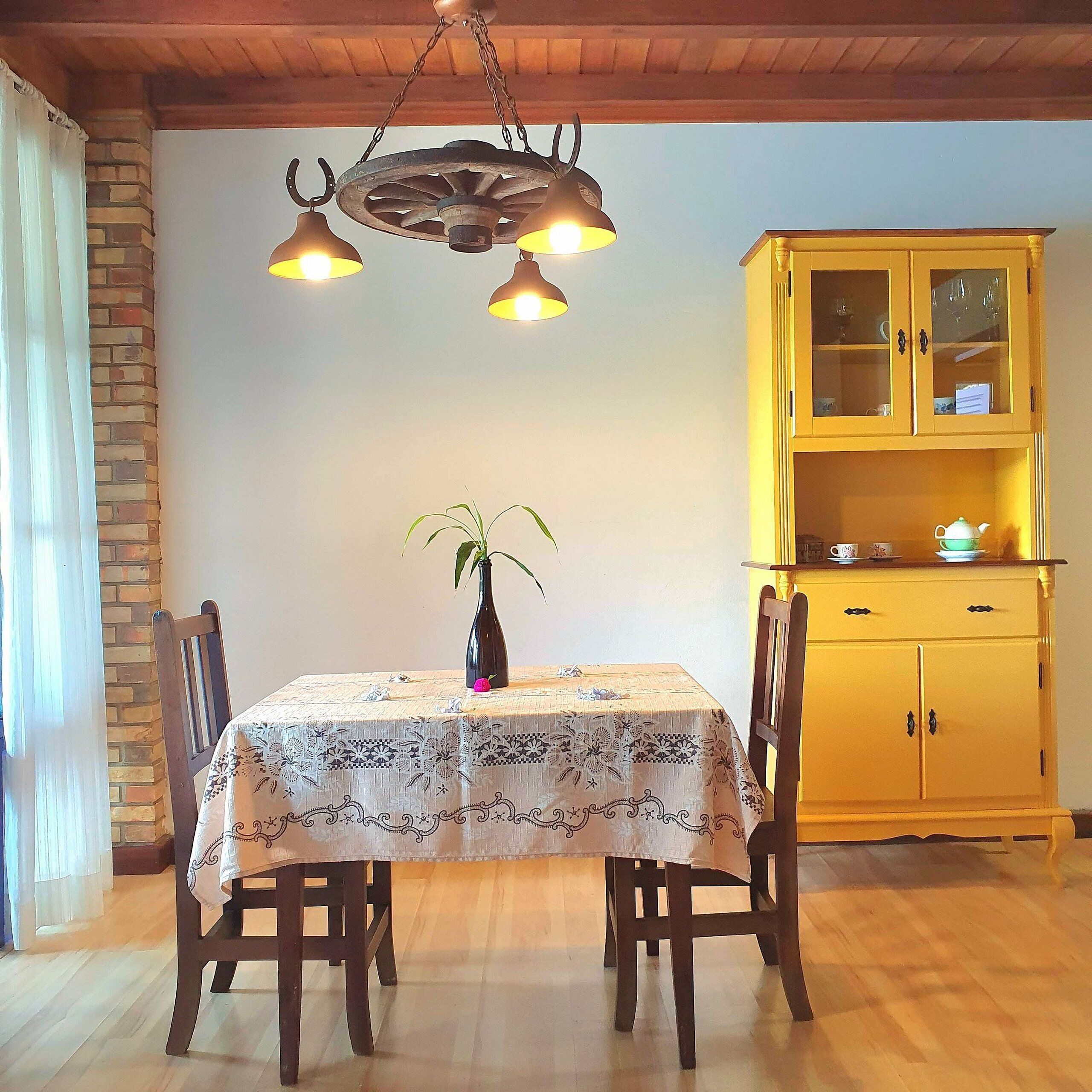Een eethoek met een tafel voor twee personen, een vaas met bloemen en een geel kastje met glazen deuren.