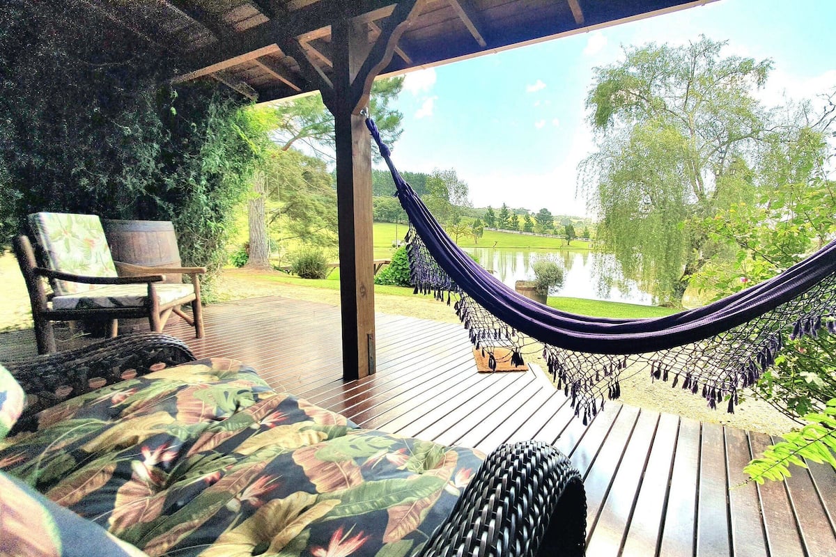 Un hamac sous une pergola en bois, une terrasse en bois, un canapé d’extérieur confortable et une vue sur un lac entouré d’arbres.