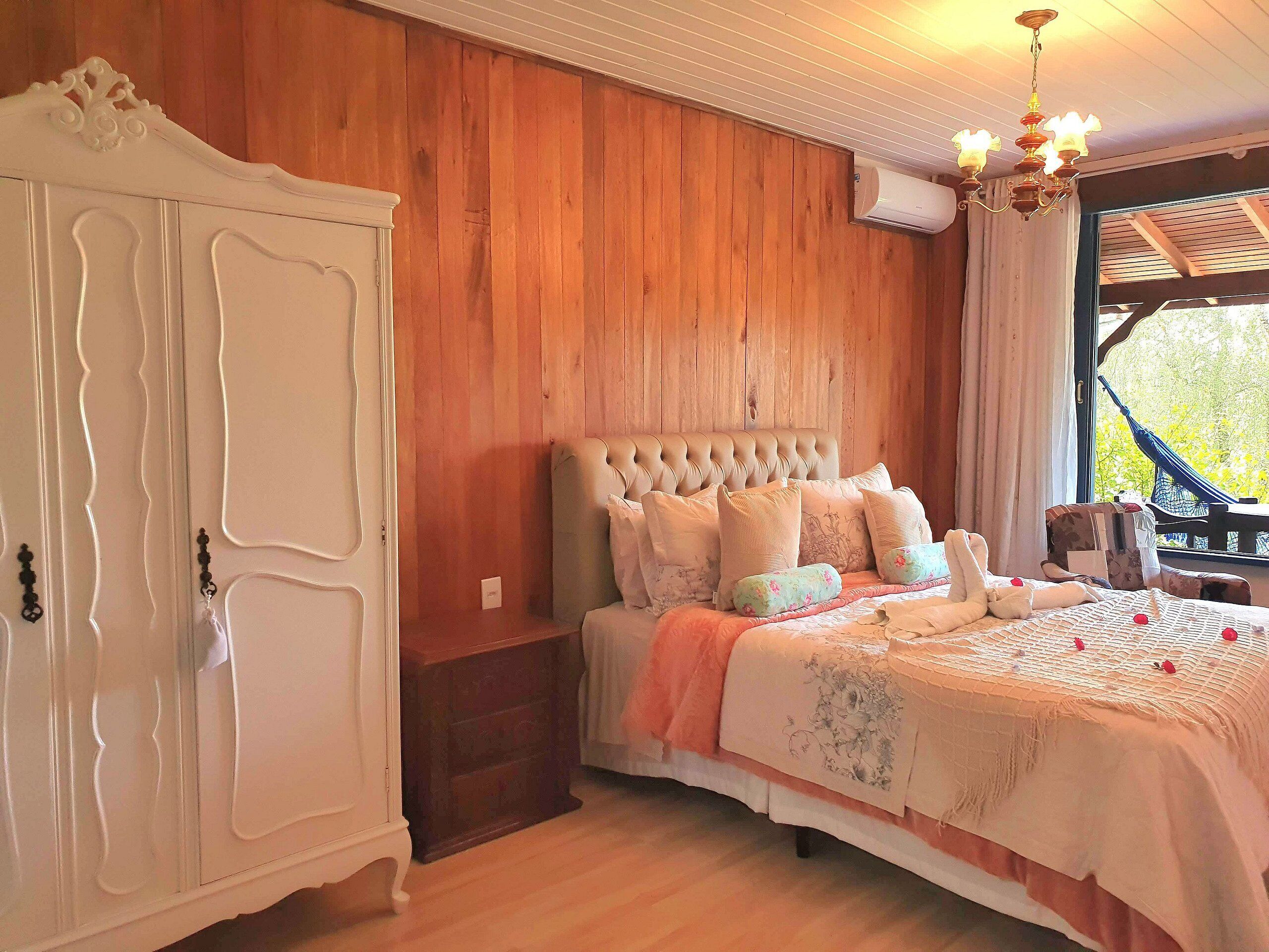 Ein Schlafzimmer mit einer holzverkleideten Wand, einem Bett mit einem gesteppten Kopfteil, einem weißen Kleiderschrank und einem Fenster mit Blick auf Grünflächen.