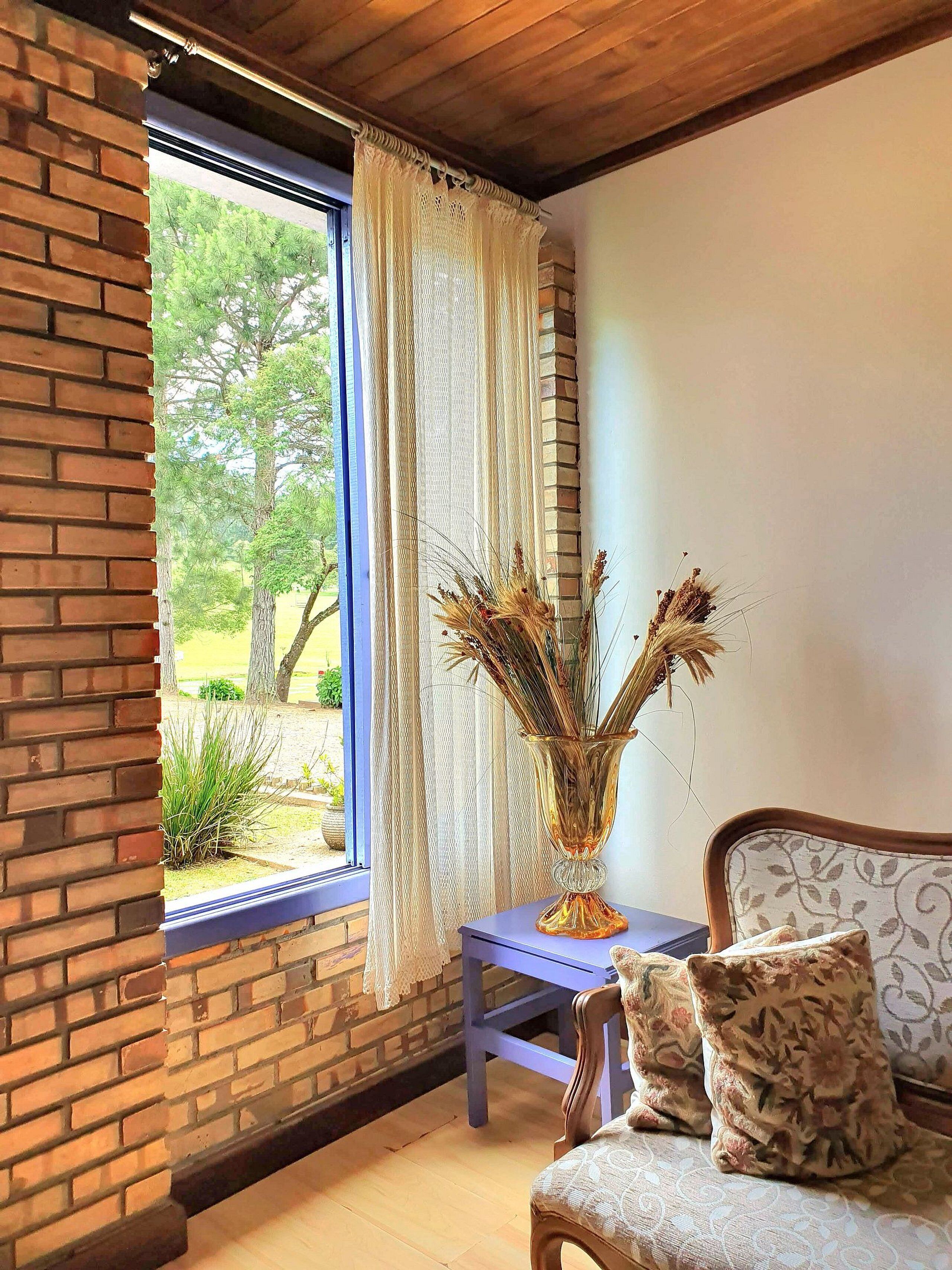 A room with a brick wall, a window with blue blinds, a wooden ceiling, a blue table, and a chair with floral cushions.