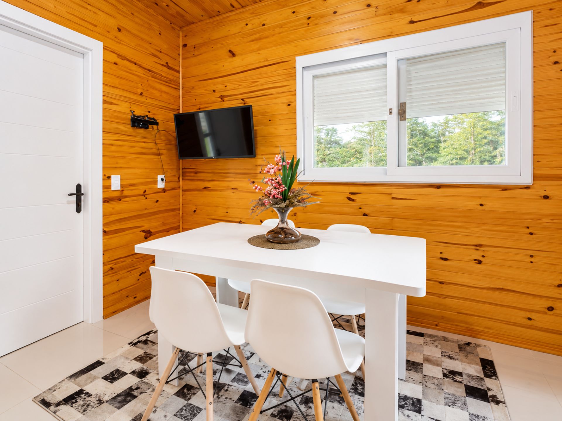 Ein Zimmer mit Holzw änden, einer weißen Tür, einem weißen Tisch mit zwei Stühlen, einem an der Wand montierten Flachbildfernseher und einem Fenster mit Jalousien.