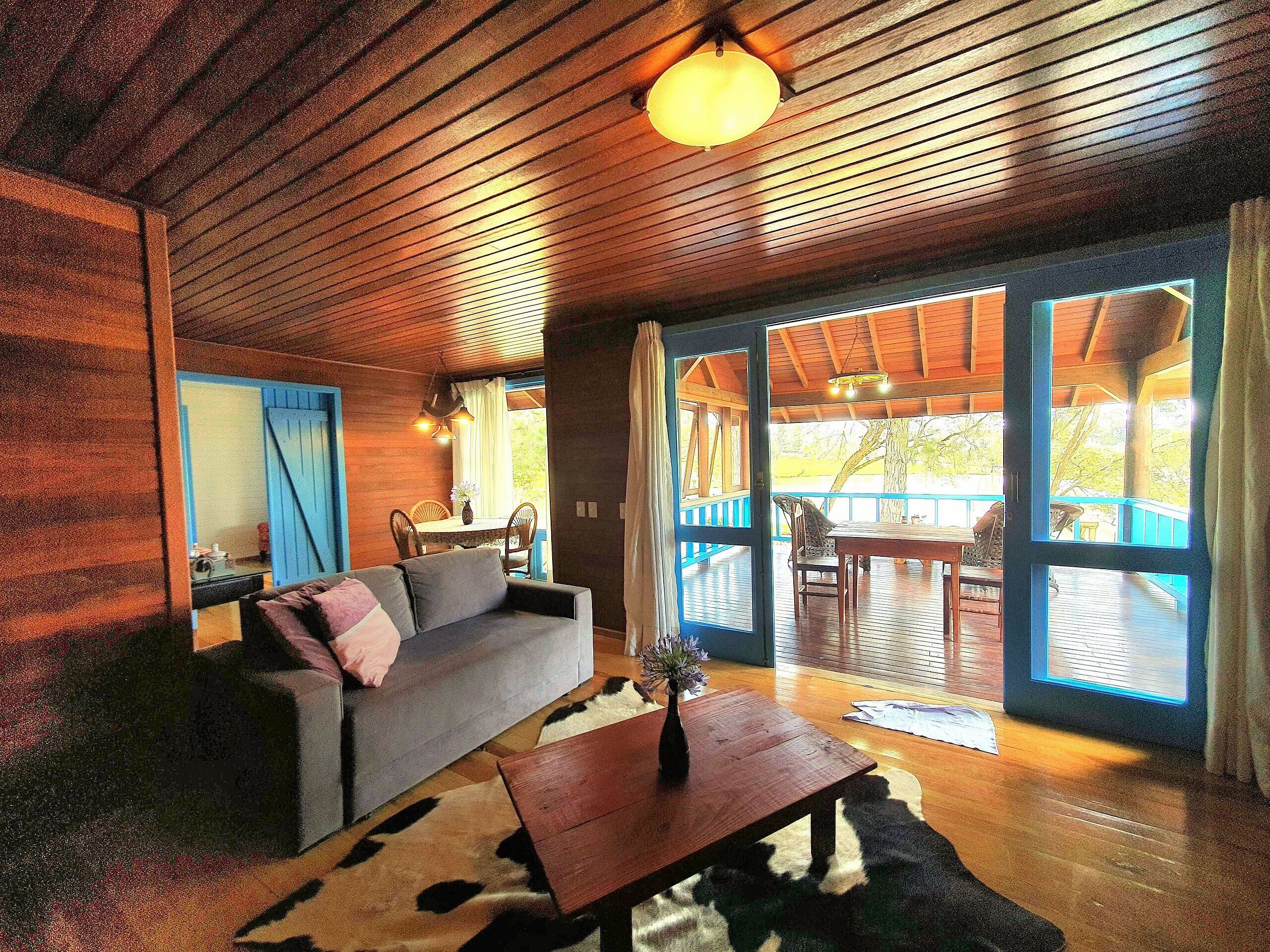 A cozy living room with a sofa, a coffee table, and a rug. There is a dining area with a table and chairs. The room has wooden walls and ceiling, and large windows.