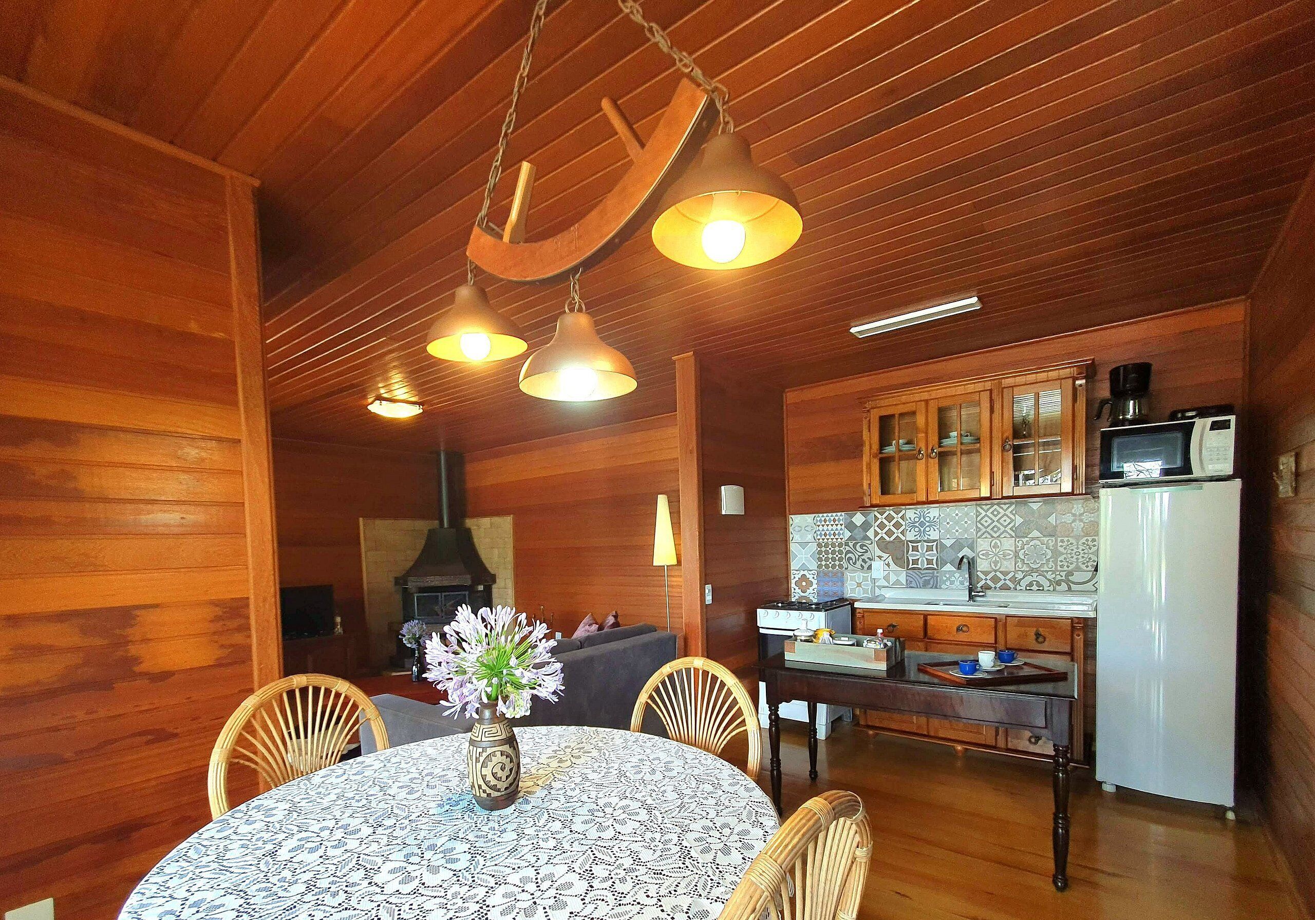 A wooden interior with a dining table, chairs, a vase with flowers, and a kitchen area with cabinets and appliances.