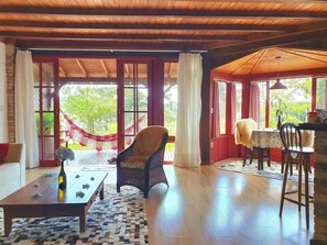 A cozy room with a wooden ceiling, a hammock, a table with a bottle, and a view of greenery outside. - Rancho das Neves - Rancho Queimado - SC (Rancho Queimado)