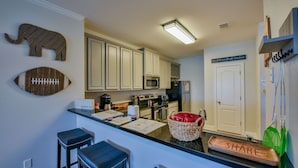 A kitchen with white cabinets, a microwave, and a refrigerator. There are two stools and a basket with red items. A sign on the wall says "share". - Welcome to Tusk-Away! (Tuscaloosa)