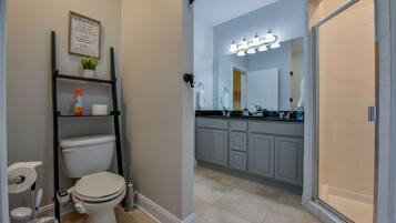 A bathroom with a toilet, sink, and vanity. There is a shelf with toiletries and a framed sign on the wall.