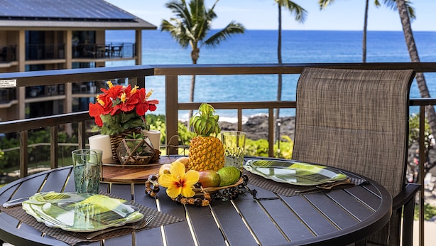 A round table with a tropical fruit arrangement, a glass of water, and a candle on a balcony overlooking the ocean with palm trees.
