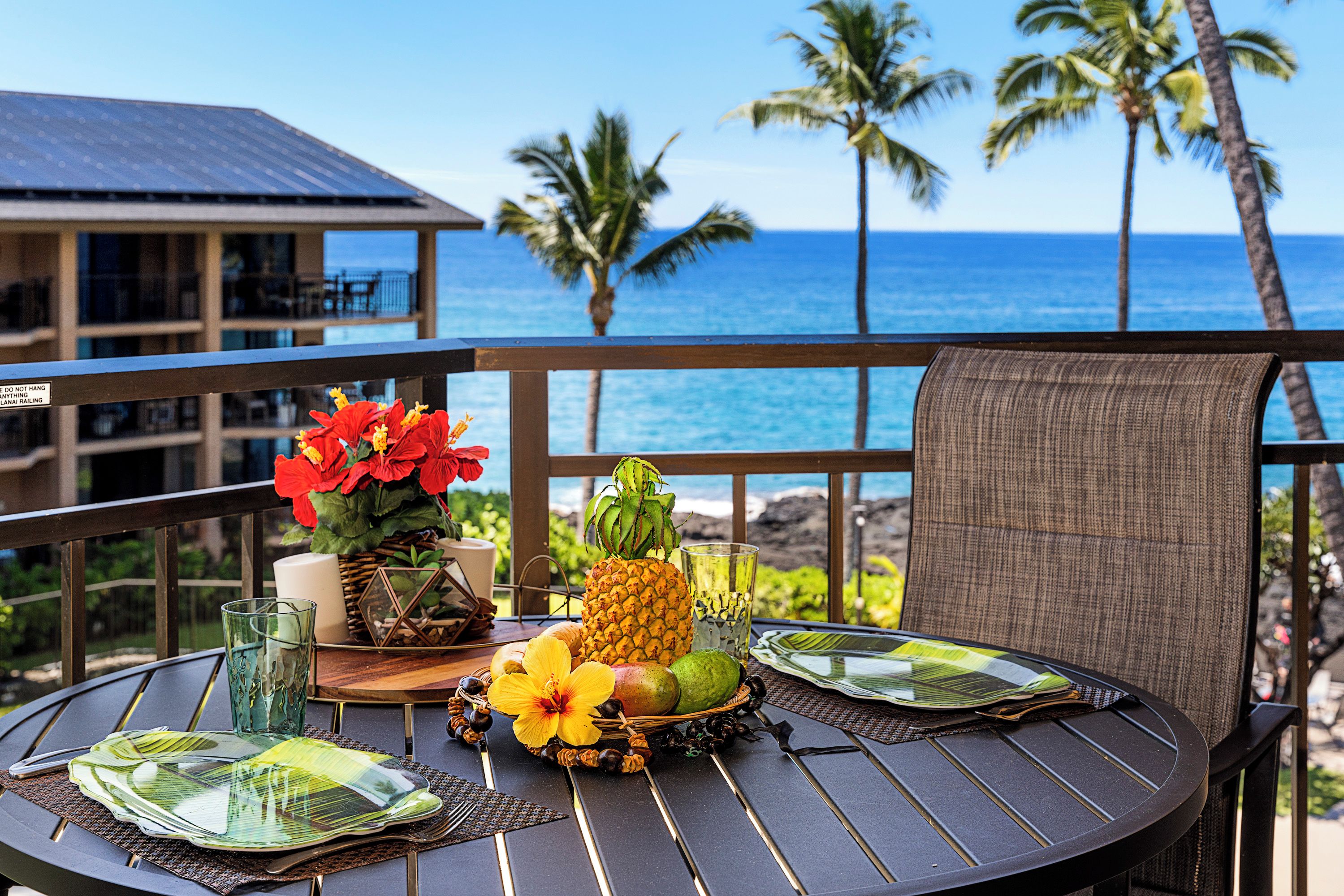 A round table with a tropical fruit arrangement, a glass of water, and a candle on a balcony overlooking the ocean with palm trees.