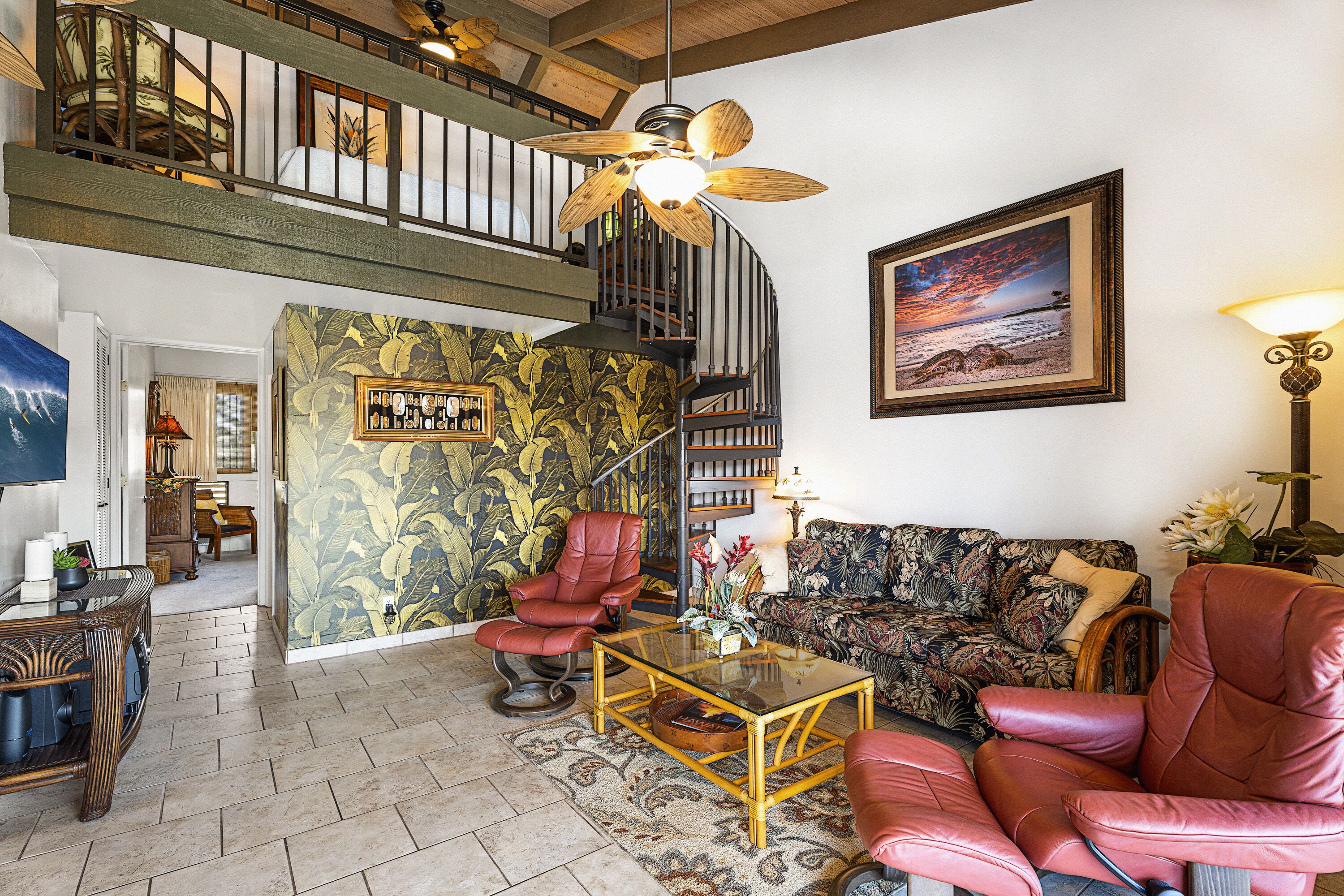 A spacious living room with a high ceiling, a staircase, a large window with a view, and a tropical wallpaper design.