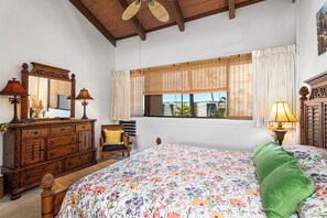 A bedroom with a floral bedspread, wooden dresser, and a large window with blinds.