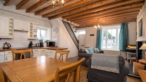 Une cuisine chaleureuse et bien éclairée, avec des poutres en bois, un coin repas comprenant une table et des chaises en bois, un salon avec un canapé et un téléviseur, et un escalier menant à un autre niveau.