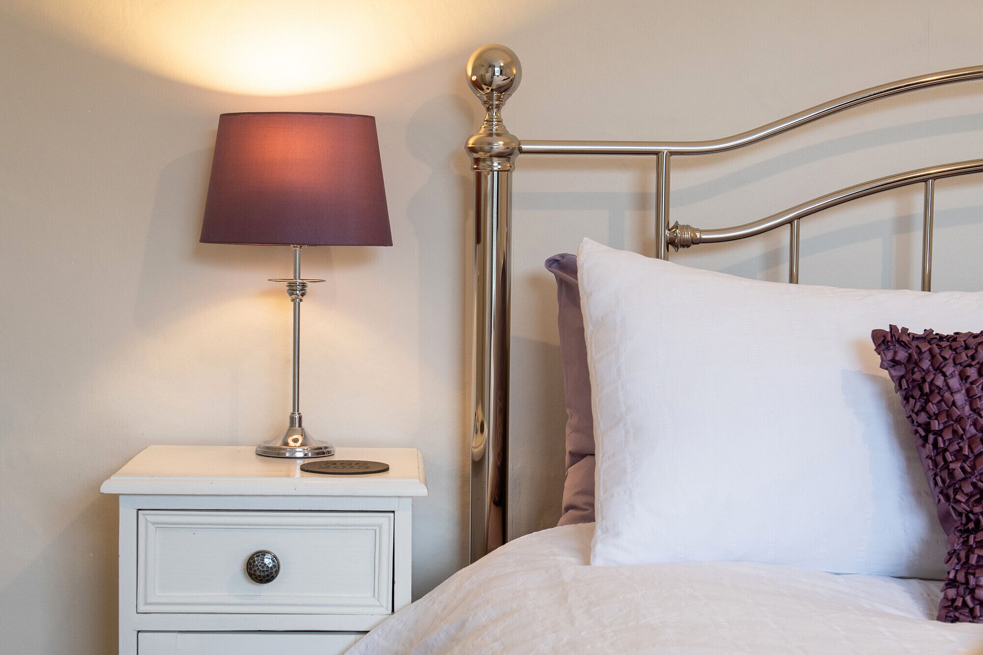 A neatly made bed with a white pillow and a decorative pillow on a white nightstand with a lamp.