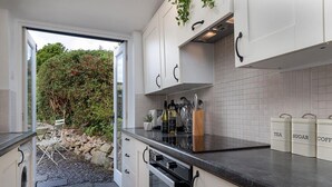 A modern kitchen with white cabinets, a black countertop, and a view of a garden through an open door. - Red Robin Cottage - One Bedroom Cottage, Sleeps 2 (Brynsiencyn)