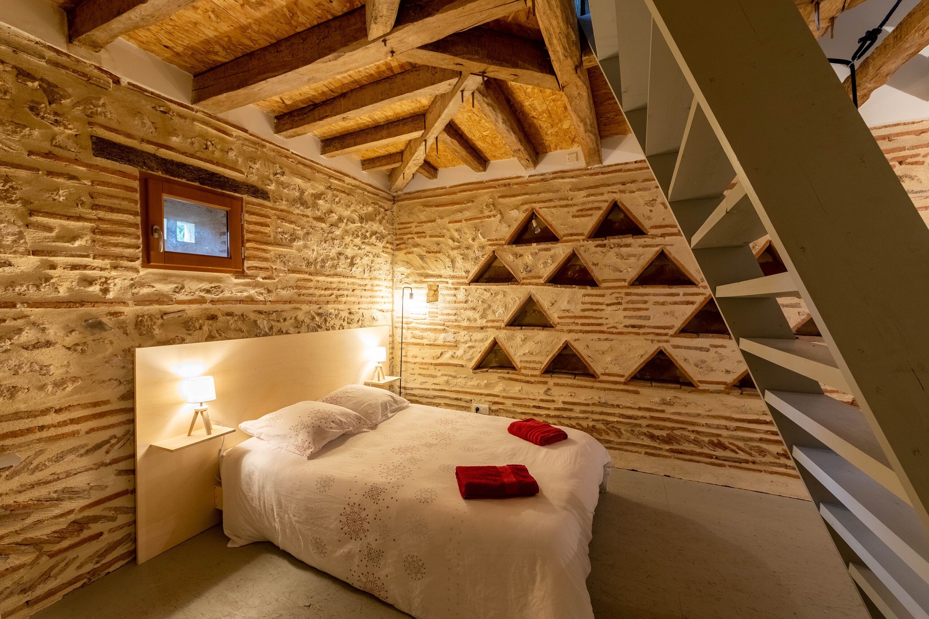 A cozy bedroom with a stone wall, a wooden headboard, a bed with white bedding, a nightstand with a lamp, and a wooden staircase.