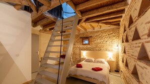 A bedroom with a wooden ceiling, a bed with white bedding and red pillows, a nightstand with a lamp, and a small window with a picture.
