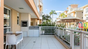 A balcony with a table and chairs, a grill, and a view of neighboring buildings and palm trees. - 350m from the Beach | Pool | Barbecue #IA20 (Florianópolis)