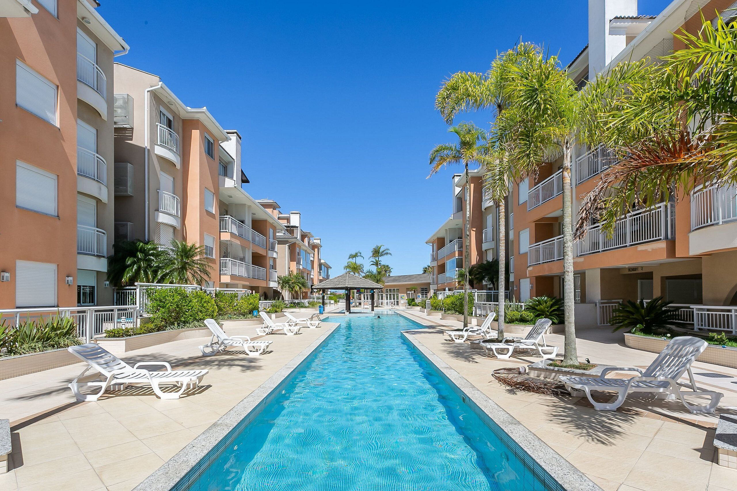 A swimming pool with lounge chairs, surrounded by multi-story buildings with balconies and palm trees.