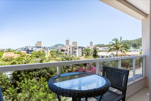 Varanda com mesa de vidro e cadeiras de vime, com vista para uma área residencial com casas e vegetação.