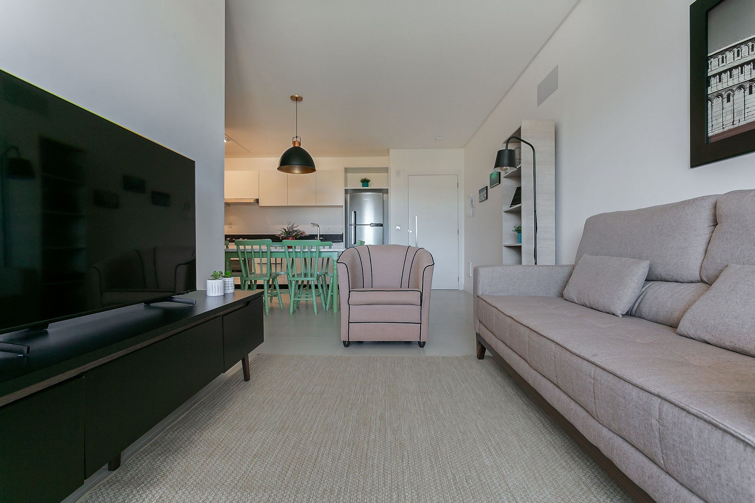 Ein modernes Wohnzimmer mit einem Flachbildfernseher, einer Couch und einem Sessel. Im Hintergrund ist eine Küche mit Esstisch und Stühlen zu sehen.