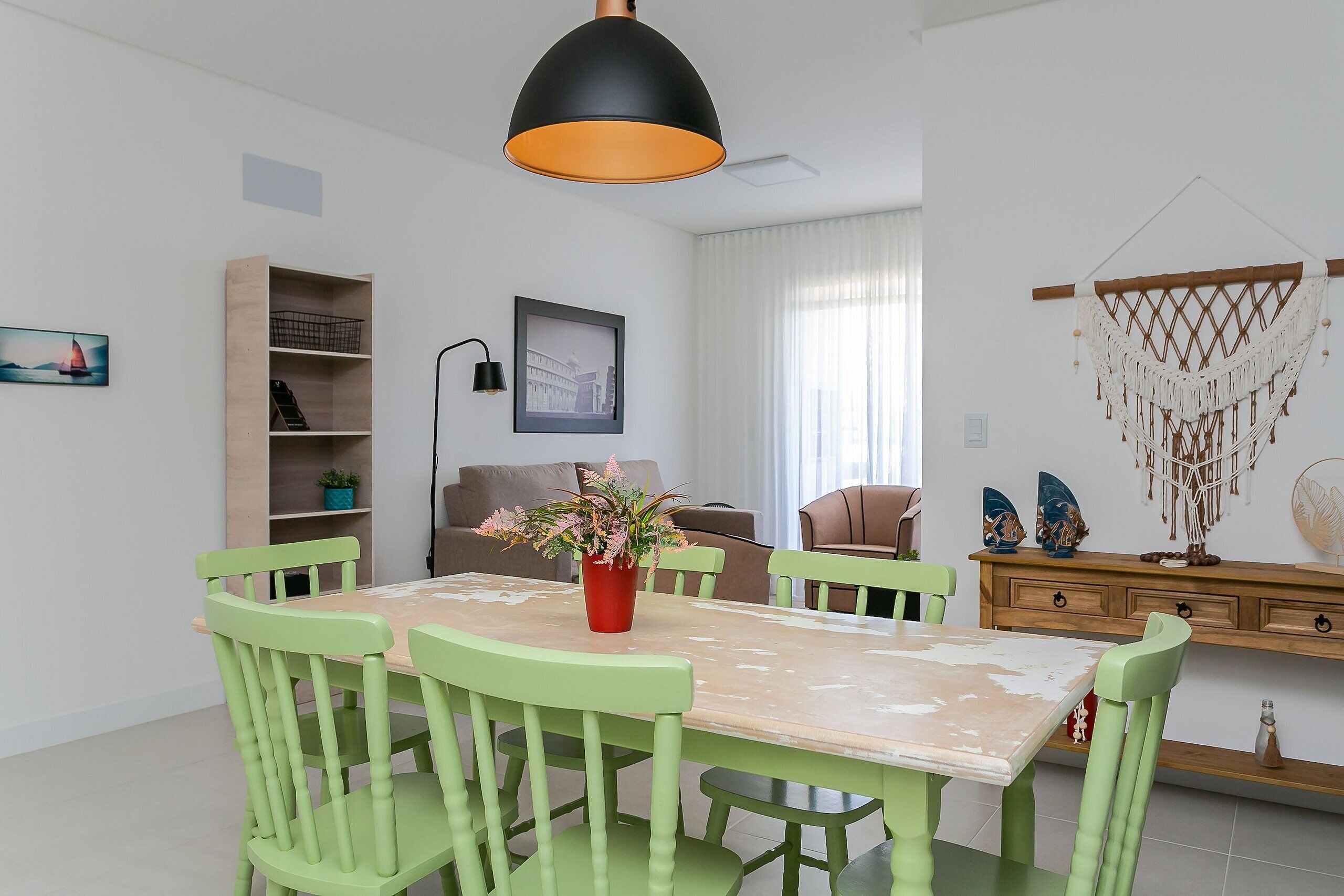 Un coin repas comprenant une table, des chaises, une suspension, une bibliothèque, un tableau encadré et une décoration murale en macramé.